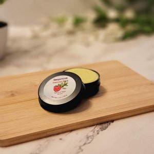 May include: Two small black tins of cuticle balm on a wooden cutting board. The tin on top has a label that reads "Supernatural Cuticle Balm Apple & Sage Scent Net Wt 1 oz / 28 g".
