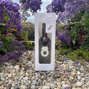 Puede incluir: Una bolsa de regalo blanca con una botella de vino dentro. La bolsa tiene una etiqueta que dice "¿Quieres ser mi dama de honor?" con un par de tacones altos dibujados en ella. La botella tiene un anillo blanco con perlas alrededor.