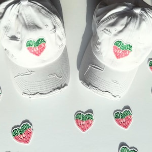 May include: Two white baseball caps with distressed details, each featuring a heart-shaped patch with the text "GO BIRDS" in green and red. Several matching heart patches are scattered around the caps.