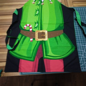 May include: Green and black apron with an elf costume design. The apron features a green tunic, red pants, a brown belt with a gold buckle, and a white collar. A candy cane is also visible on the apron.