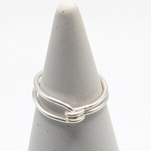 May include: A silver-colored ring with a double-band design. The ring is displayed on a gray, cone-shaped stand against a white background. The ring's bands are connected with a wrapped detail.