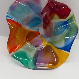 May include: A colorful, freeform glass bowl with a wavy edge. The bowl features a vibrant mix of colors, including red, orange, yellow, green, blue, and purple. The glass appears translucent, allowing light to pass through and highlight the colors.
