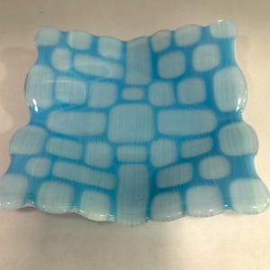 Peut inclure: Un plat en verre bleu clair avec un motif texturé en forme de briques. Les bords du plat sont ondulés et la forme générale est rectangulaire. La couleur est un bleu pâle et doux avec des accents blancs.