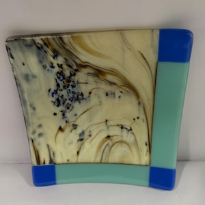 May include: A decorative glass piece with a unique design. The main section features swirling patterns in cream, brown, and black, with scattered blue accents. A light teal border and blue corner accents complete the design.