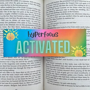 Op de afbeelding: Een kleurrijke bladwijzer met de woorden "hyperfocus ACTIVATED" in grote letters. De bladwijzer, met een gradiënt achtergrond en hersengrafieken, ligt in een open boek.