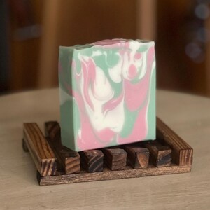 Puede incluir: Una pastilla de jabón artesanal con remolinos rosas, blancos y verdes, colocada sobre un pequeño jabonera de madera oscura. El jabón tiene un aspecto veteado y la jabonera tiene un diseño de listones.