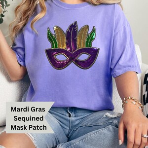 May include: A purple t-shirt with a sequined Mardi Gras mask patch. The mask is purple, green, and gold. The text "Mardi Gras Sequined Mask Patch" is written below the mask.