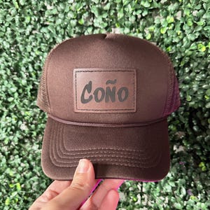 May include: A brown trucker hat with a mesh back and a curved brim. The hat features a rectangular patch with the word "COÑO" in black lettering. The hat is made of a soft material and has a brown rope detail.