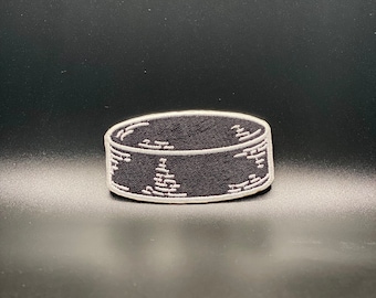 May include: A black and white embroidered patch of a hockey puck. The puck is outlined in white and has a black centre.