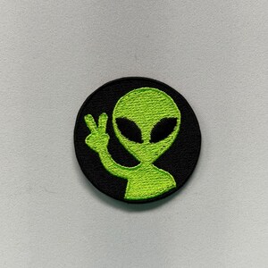 May include: A circular embroidered patch featuring a bright lime green alien figure against a solid black background. The alien is depicted from the chest up, making a peace sign gesture with its hand, and has large, dark, almond-shaped eyes.