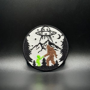 Peut inclure: Un patch rond brodé avec un fond blanc représentant un extraterrestre vert et un Bigfoot brun debout devant une montagne avec un OVNI au-dessus d'eux. Le patch a une bordure noire et est entouré d'un fond noir.