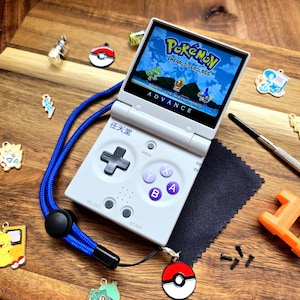 May include: A white Game Boy Advance SP handheld console with a blue lanyard, displaying a Pok&eacute;mon game. The image includes various Pok&eacute;mon-themed charms and a cleaning cloth, set on a wooden surface.