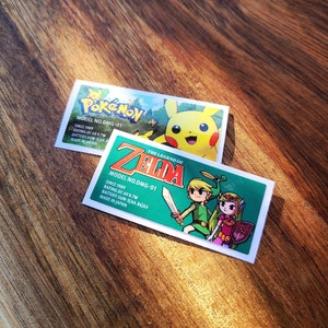 Puede incluir: Dos etiquetas de cartucho de juego para la Nintendo Game Boy. La etiqueta superior es para Pokemon y presenta un Pikachu amarillo. La etiqueta inferior es para The Legend of Zelda y presenta a Link y la princesa Zelda. Ambas etiquetas tienen el número de modelo DMG-01, el año 1989 y el texto "Hecho en Japón".