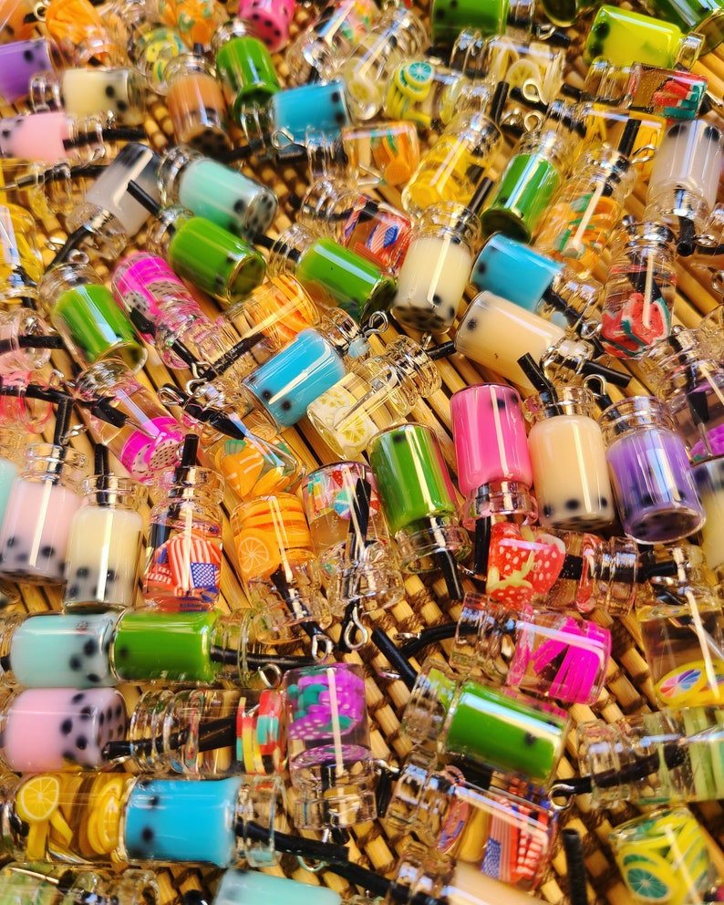 May include: A collection of miniature, colorful bubble tea charms. The charms feature various colors and flavors, including pink, green, blue, and yellow, with black tapioca pearls. Each charm has a small straw and a silver-colored cap.