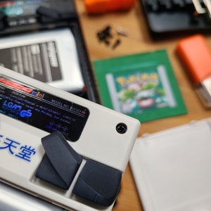 Puede incluir: Un dispositivo de juego port&aacute;til blanco con botones negros y una etiqueta azul y blanca que dice "LGADGS" y "天堂". La etiqueta tambi&eacute;n incluye el texto "MODEL NO. MIYOO" y "ADAPTER INPUT USB-C 5V".
