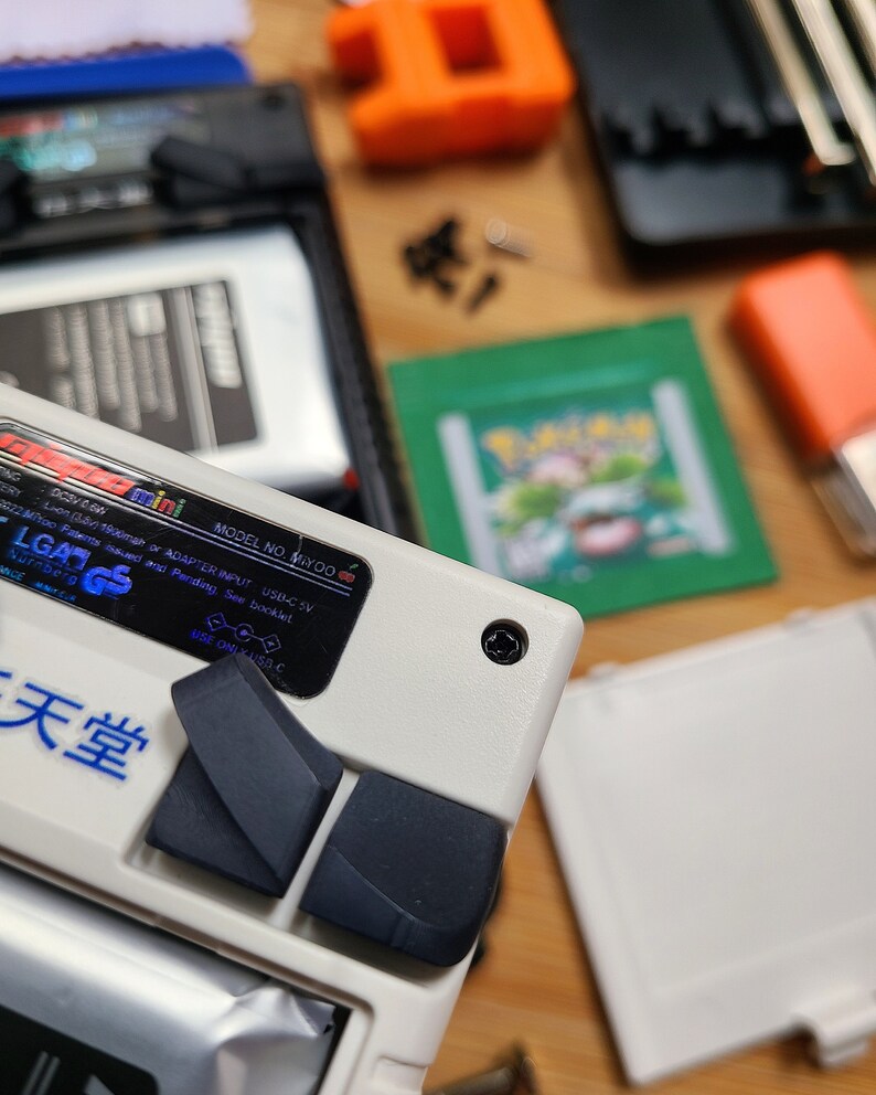 Puede incluir: Un dispositivo de juego port&aacute;til blanco con botones negros y una etiqueta azul y blanca que dice "LGADGS" y "天堂". La etiqueta tambi&eacute;n incluye el texto "MODEL NO. MIYOO" y "ADAPTER INPUT USB-C 5V".