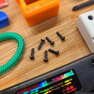 Puede incluir: Primer plano de seis peque&ntilde;os tornillos negros sobre una superficie de madera. Los tornillos est&aacute;n dispersos alrededor de un cable verde y un dispositivo electr&oacute;nico negro con una pegatina hologr&aacute;fica arco&iacute;ris que dice "mini".