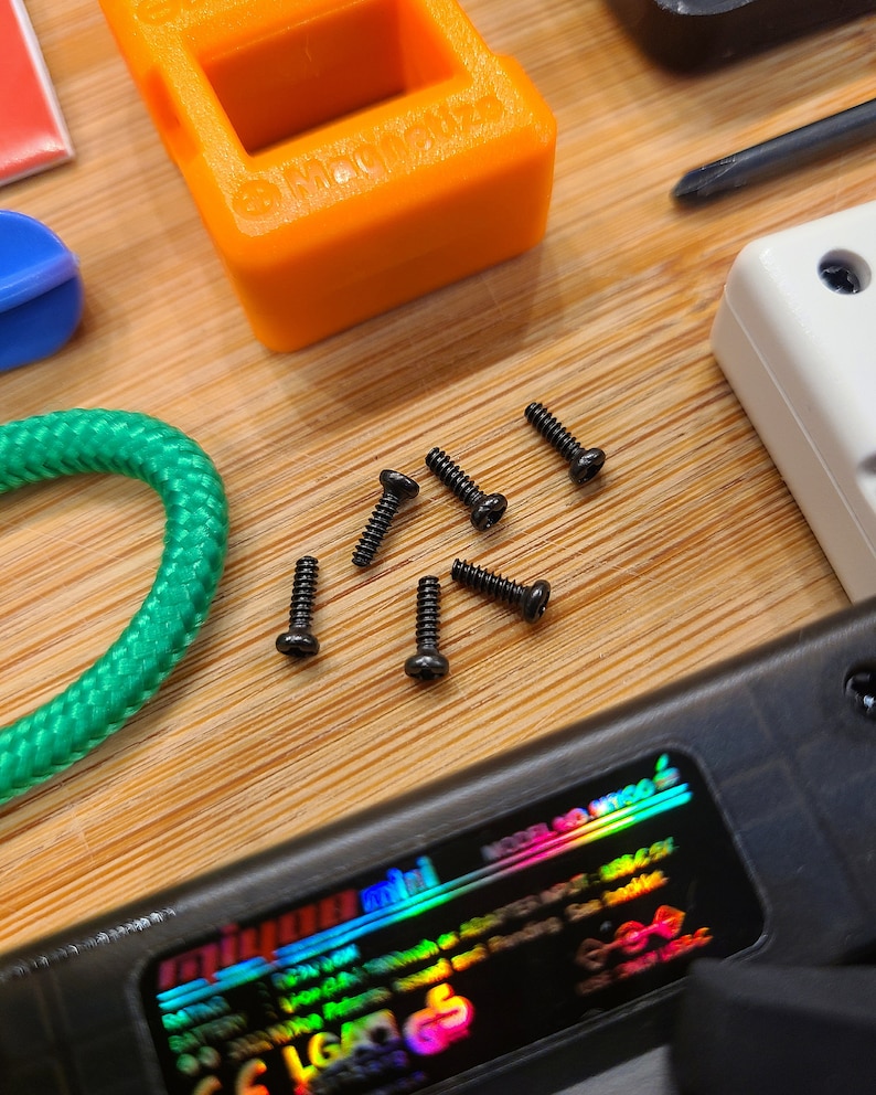 Puede incluir: Primer plano de seis peque&ntilde;os tornillos negros sobre una superficie de madera. Los tornillos est&aacute;n dispersos alrededor de un cable verde y un dispositivo electr&oacute;nico negro con una pegatina hologr&aacute;fica arco&iacute;ris que dice "mini".