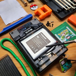 Puede incluir: Una consola de juegos port&aacute;til desmontada con un cable verde, una herramienta de palanca azul, una peque&ntilde;a pieza de pl&aacute;stico naranja, un pa&ntilde;o de limpieza blanco, una carta de Pokemon en blanco y negro, un peque&ntilde;o pin de Pokemon negro y rojo, un peque&ntilde;o amuleto de Pokemon negro y verde, un juego de destornilladores plateados, un destornillador plateado, una bater&iacute;a negra y plateada, y una punta de destornillador negra y plateada.