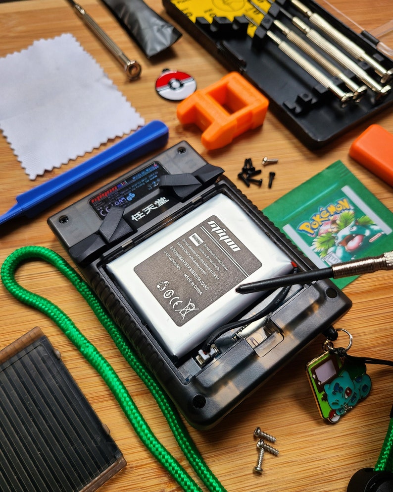 Puede incluir: Una consola de juegos port&aacute;til desmontada con un cable verde, una herramienta de palanca azul, una peque&ntilde;a pieza de pl&aacute;stico naranja, un pa&ntilde;o de limpieza blanco, una carta de Pokemon en blanco y negro, un peque&ntilde;o pin de Pokemon negro y rojo, un peque&ntilde;o amuleto de Pokemon negro y verde, un juego de destornilladores plateados, un destornillador plateado, una bater&iacute;a negra y plateada, y una punta de destornillador negra y plateada.