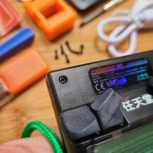 Puede incluir: Un dispositivo electr&oacute;nico de mano negro con un cable verde conectado. El dispositivo tiene un peque&ntilde;o bot&oacute;n negro en la parte superior y un bot&oacute;n negro m&aacute;s grande en el lateral. El dispositivo tiene una pantalla con un fondo verde y texto blanco. El texto en la pantalla dice "任天堂".