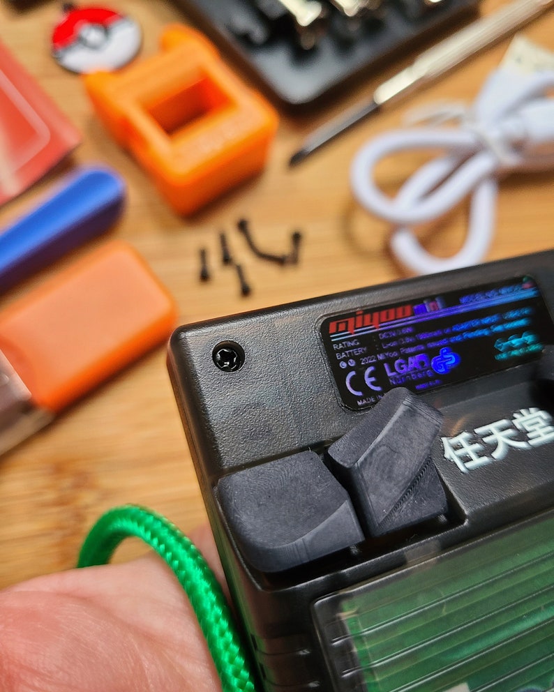 Puede incluir: Un dispositivo electr&oacute;nico de mano negro con un cable verde conectado. El dispositivo tiene un peque&ntilde;o bot&oacute;n negro en la parte superior y un bot&oacute;n negro m&aacute;s grande en el lateral. El dispositivo tiene una pantalla con un fondo verde y texto blanco. El texto en la pantalla dice "任天堂".