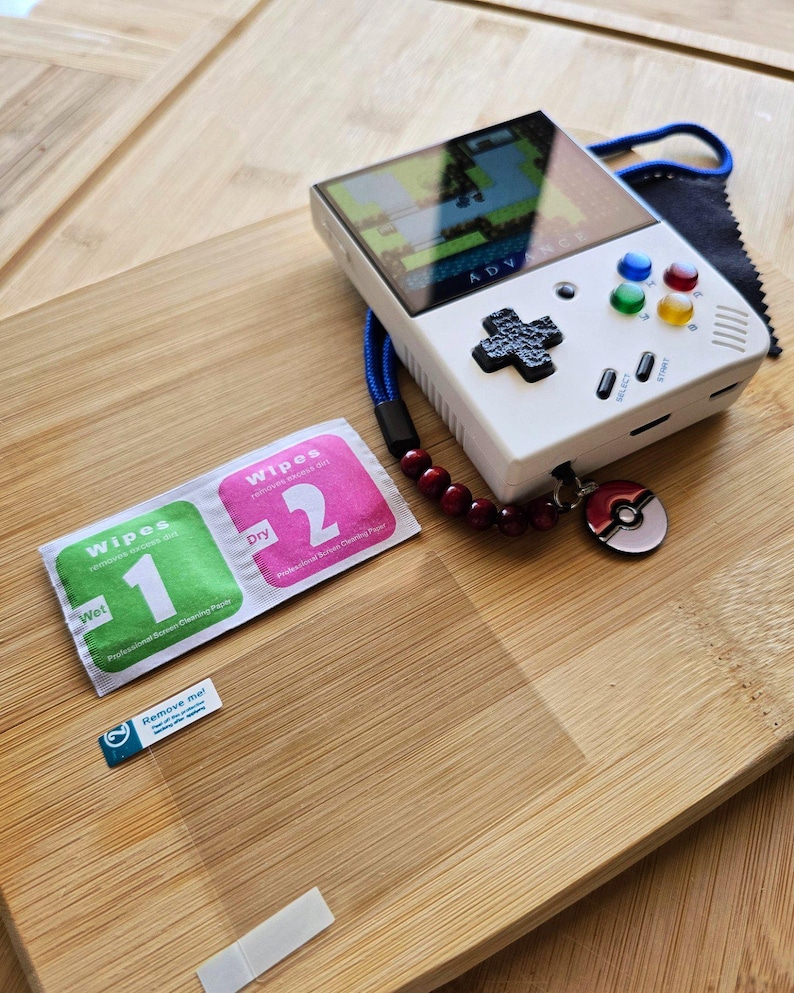 May include: A white handheld gaming console with a screen displaying a game. It has colored buttons, a directional pad, and a Pokemon charm. Accessories include screen cleaning wipes and a screen protector on a wooden surface.