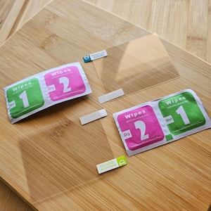 May include: A clear screen protector kit with two cleaning wipes and adhesive strips, displayed on a wooden surface. The wipes are labeled "Wet" and "Dry" with numbers 1 and 2. The kit also includes a "Remove me" sticker.