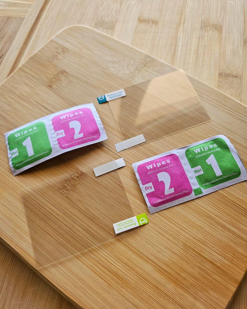 May include: A clear screen protector kit with two cleaning wipes and adhesive strips, displayed on a wooden surface. The wipes are labeled "Wet" and "Dry" with numbers 1 and 2. The kit also includes a "Remove me" sticker.