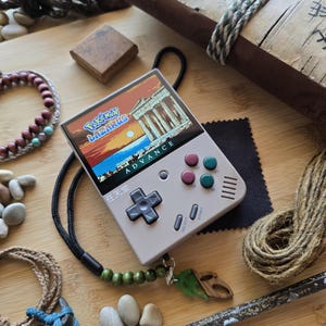 May include: A handheld gaming console with a screen displaying a video game scene. The console is light gray with colored buttons and the text "Pokemon Lazarus Advance". A black lanyard with wooden beads and a leaf pendant is attached.