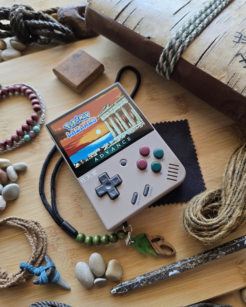 May include: A handheld gaming console with a screen displaying a video game scene. The console is light gray with colored buttons and the text "Pokemon Lazarus Advance". A black lanyard with wooden beads and a leaf pendant is attached.
