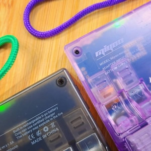 Puede incluir: Primer plano de un dispositivo de juego port&aacute;til negro y morado con el texto "Miyoo-mini" y "MODEL: Miyoo-mini" impreso en el dispositivo. El dispositivo tiene una bater&iacute;a Li-Po recargable incorporada y un cargador certificado. El texto "Built in rechargeable li-po battery" y "Only can use certified charger" est&aacute; impreso en el dispositivo. El texto "The battery may explode in the fire" est&aacute; impreso en el dispositivo con un s&iacute;mbolo de advertencia. El texto "CE" y "MADE IN CHINA" est&aacute; impreso en el dispositivo.