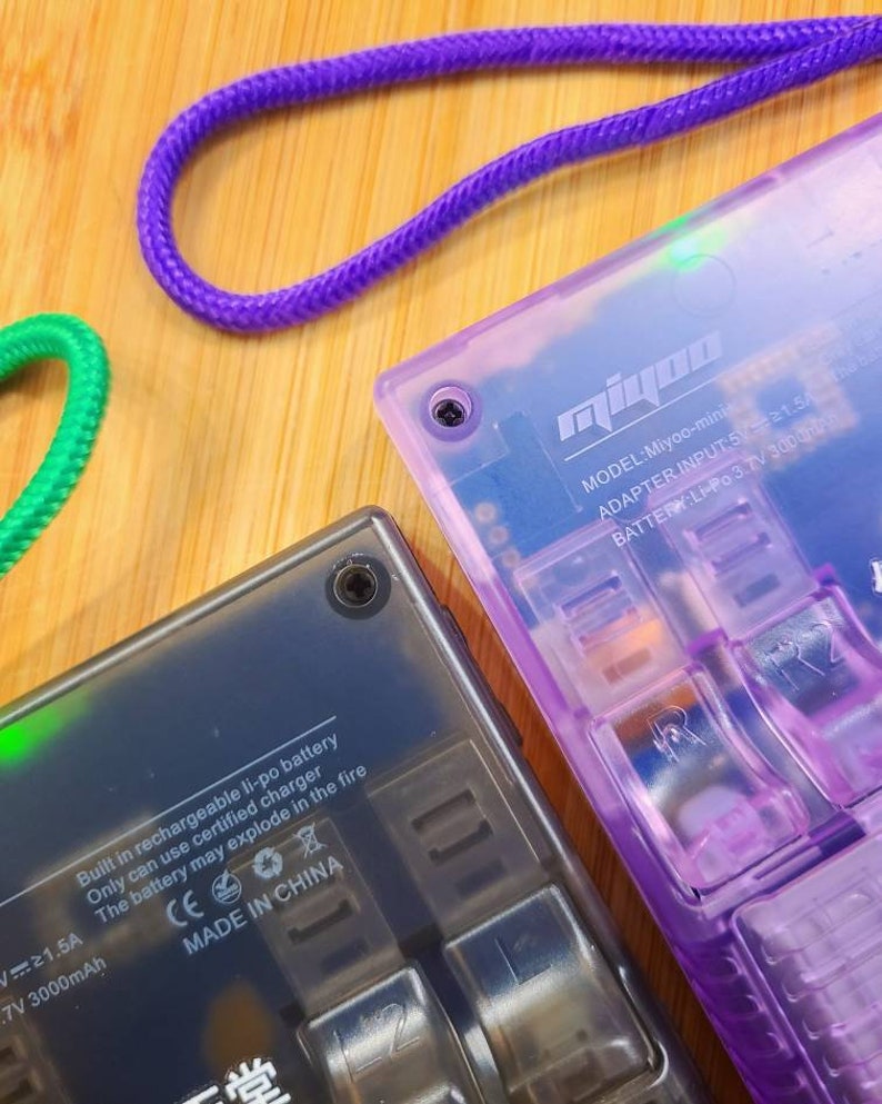Puede incluir: Primer plano de un dispositivo de juego port&aacute;til negro y morado con el texto "Miyoo-mini" y "MODEL: Miyoo-mini" impreso en el dispositivo. El dispositivo tiene una bater&iacute;a Li-Po recargable incorporada y un cargador certificado. El texto "Built in rechargeable li-po battery" y "Only can use certified charger" est&aacute; impreso en el dispositivo. El texto "The battery may explode in the fire" est&aacute; impreso en el dispositivo con un s&iacute;mbolo de advertencia. El texto "CE" y "MADE IN CHINA" est&aacute; impreso en el dispositivo.