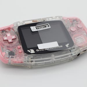 May include: A clear pink handheld game console with a black screen and buttons. The console has a sticker on the screen that says "FRONT 00". The console also has the text "GAME BOY ADVANCE" printed on the front.