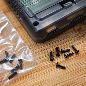 Puede incluir: Primer plano de una caja de pl&aacute;stico negro con un puerto USB-C y un peque&ntilde;o bot&oacute;n. La caja est&aacute; rodeada de un mont&oacute;n de peque&ntilde;os tornillos negros.