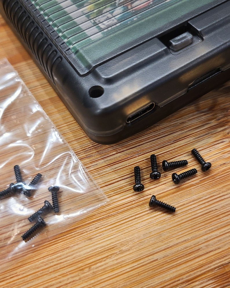 Puede incluir: Primer plano de una caja de pl&aacute;stico negro con un puerto USB-C y un peque&ntilde;o bot&oacute;n. La caja est&aacute; rodeada de un mont&oacute;n de peque&ntilde;os tornillos negros.