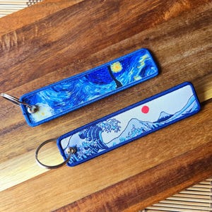 Puede incluir: Dos llaveros de tela azul y blanca con pinturas famosas. Un llavero tiene una imagen azul y blanca de "La Gran Ola de Kanagawa" de Hokusai. El otro llavero tiene una imagen azul y blanca de "La Noche Estrellada" de Vincent van Gogh.