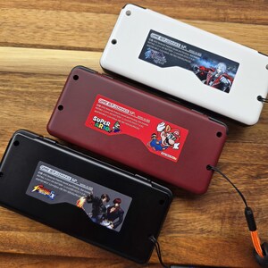 Puede incluir: Tres consolas Game Boy Advance SP con carcasas de diferentes colores. La consola superior es blanca con una pegatina de un personaje del videojuego Castlevania: Symphony of the Night. La consola del medio es roja con una pegatina de Super Mario. La consola inferior es negra con una pegatina de un personaje del videojuego The King of Fighters.
