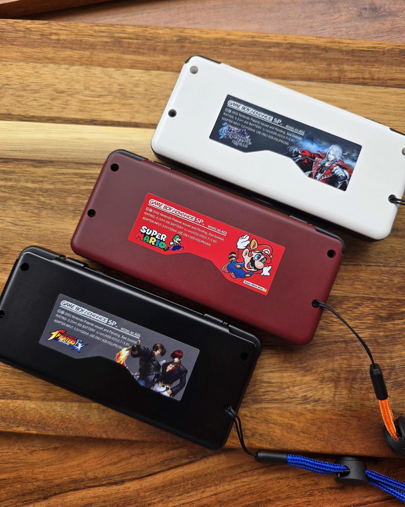 Puede incluir: Tres consolas Game Boy Advance SP con carcasas de diferentes colores. La consola superior es blanca con una pegatina de un personaje del videojuego Castlevania: Symphony of the Night. La consola del medio es roja con una pegatina de Super Mario. La consola inferior es negra con una pegatina de un personaje del videojuego The King of Fighters.