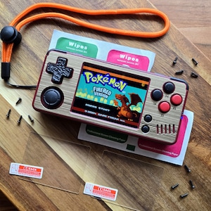 Può includere: Console di videogiochi portatile con finitura effetto legno, che mostra "Pokémon FireRed Version". Ha pulsanti rossi e neri, un D-pad e un cordino arancione. Gli accessori includono salviette e protezioni per lo schermo.