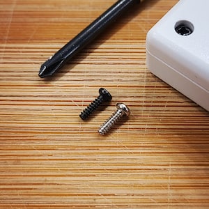 Puede incluir: Dos tornillos, uno negro y uno plateado, est&aacute;n sobre una superficie de madera. Tambi&eacute;n se puede ver un destornillador en el fondo.