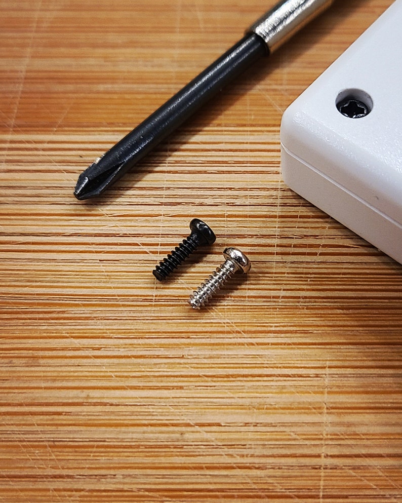 Puede incluir: Dos tornillos, uno negro y uno plateado, est&aacute;n sobre una superficie de madera. Tambi&eacute;n se puede ver un destornillador en el fondo.