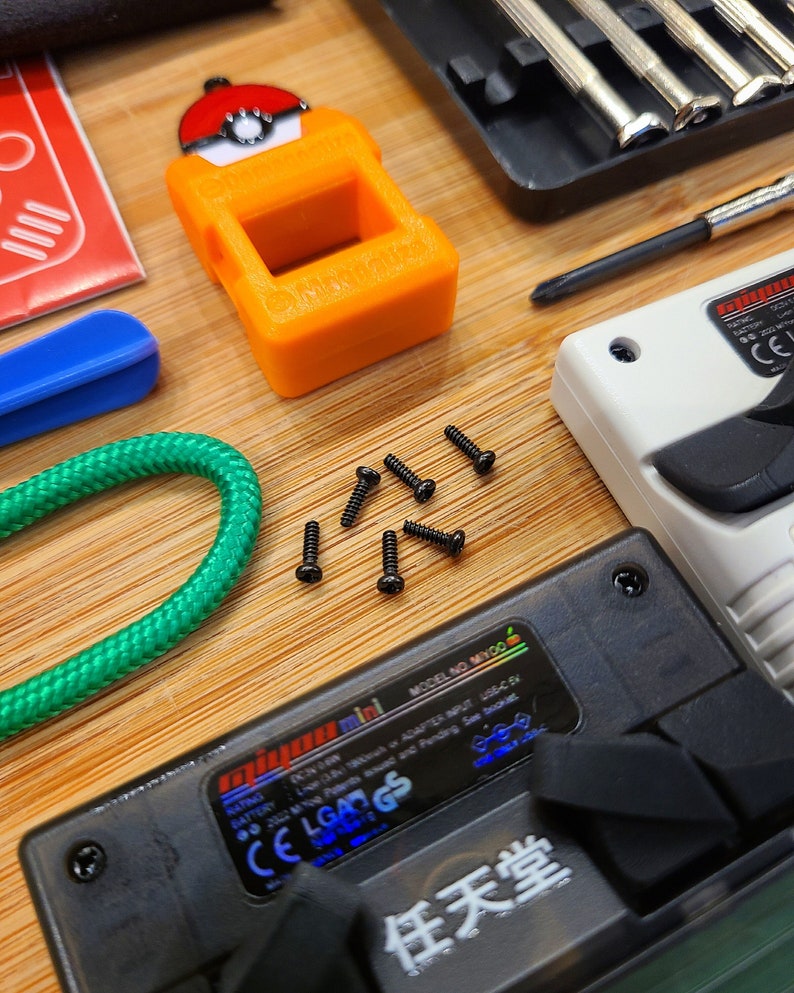 Puede incluir: Primer plano de un dispositivo electr&oacute;nico negro y azul con el texto "任天堂" en blanco. El dispositivo tiene una peque&ntilde;a pantalla y varios botones. Tambi&eacute;n hay varios tornillos peque&ntilde;os esparcidos alrededor del dispositivo.