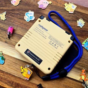 Puede incluir: Una consola de juegos port&aacute;til amarilla y negra con una correa azul. La consola tiene un logotipo de Pok&eacute;mon Center y el texto "MODEL: Miyoo-Flip, ADAPTER INPUT: 5V 1.5A, BATTERY: Li-Po 3.7V 3000mAh, Built in rechargeable li-po battery, Only can use certified charger, The battery may explode in the fire, CE, MADE IN CHINA" en la parte posterior.