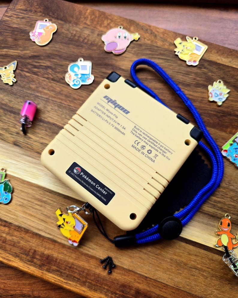 Puede incluir: Una consola de juegos port&aacute;til amarilla y negra con una correa azul. La consola tiene un logotipo de Pok&eacute;mon Center y el texto "MODEL: Miyoo-Flip, ADAPTER INPUT: 5V 1.5A, BATTERY: Li-Po 3.7V 3000mAh, Built in rechargeable li-po battery, Only can use certified charger, The battery may explode in the fire, CE, MADE IN CHINA" en la parte posterior.