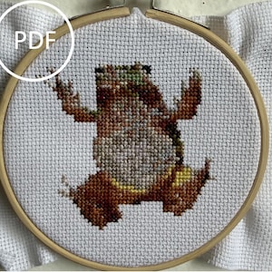 May include: A cross-stitch embroidery of a frog in a wooden hoop. The frog is brown, green, and yellow, with its arms raised. The word "PDF" is in a white circle in the upper left corner. The background is white.