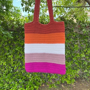 Puede incluir: Una bolsa de mano de crochet con un diseño a rayas en tonos de rojo, naranja, blanco, rosa y magenta. La bolsa tiene dos asas largas.
