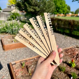May include: Wooden plant markers with the names of different vegetables written on them, including "Sweet 100s", "Yellow Peppers", "Cantaloupe", "Red Peppers", "Black Cherry Tomatoes", and "Spaghetti Squash".