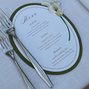 May include: A wedding menu card with a green border and floral accents, featuring a list of dishes including Caesar salad, smoked brisket, and vanilla wedding cake. Two silver forks are placed on a white linen tablecloth.