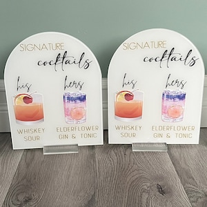May include: Four white acrylic signs with rounded tops, each displaying the text "SIGNATURE cocktails" and two cocktail options labeled "his" and "hers". The cocktails are illustrated with watercolor-style images of whiskey sour and elderflower gin and tonic.